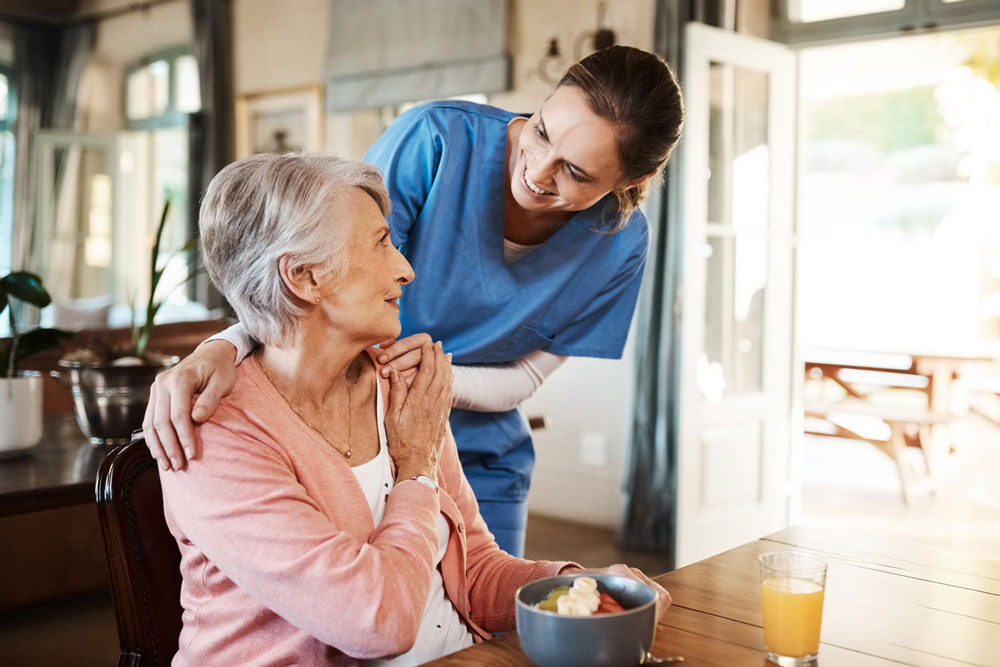 A female nurse in blue scrubs holds the shoulders of a seated older woman in a brightly lit room