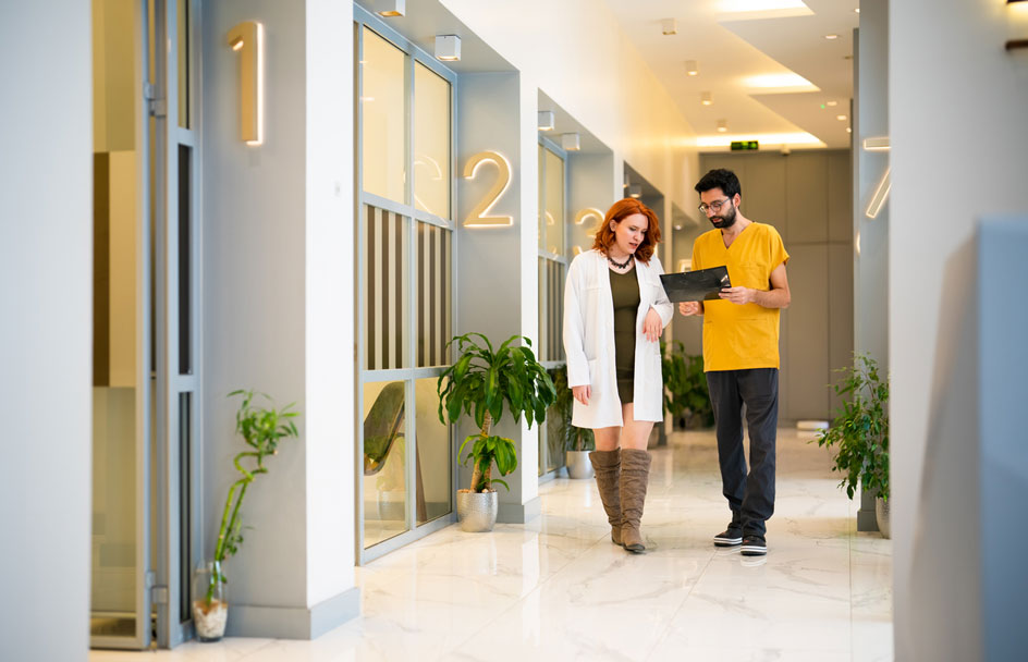 A woman in a lab coat and a man in a yellow scrub shirt walk down a bright hallway and look at a chart