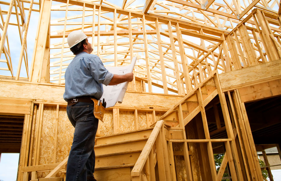 A construction worker in a hard hat holds blueprints and walks up the stairs in a new build