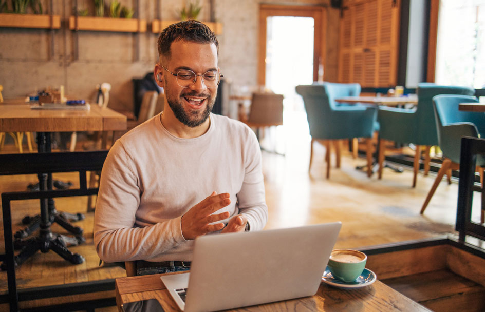 A man in glasses and earphones sits in an empty lounge and smiles at a laptop