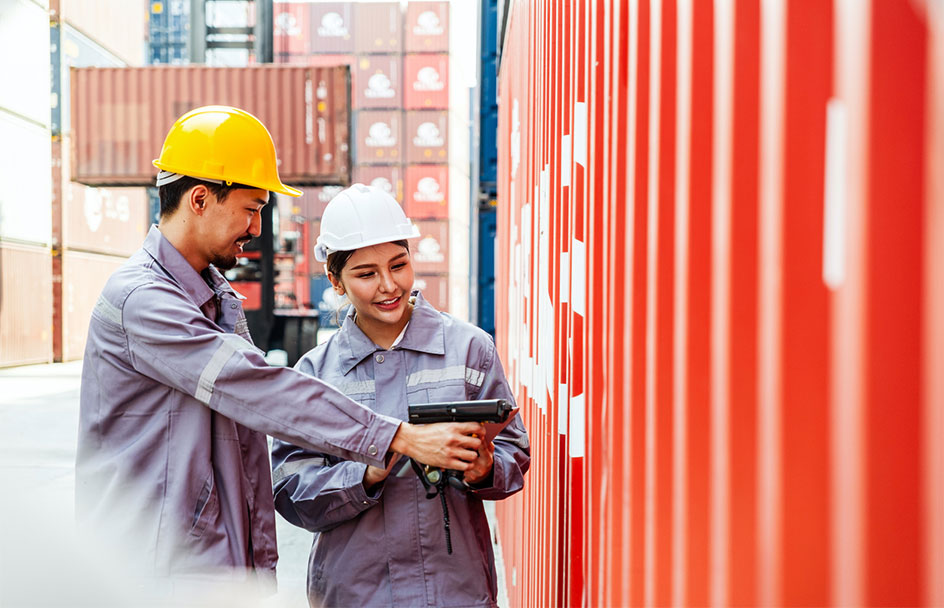 Two workers in jumpsuits and hard hats stand beside a cargo container