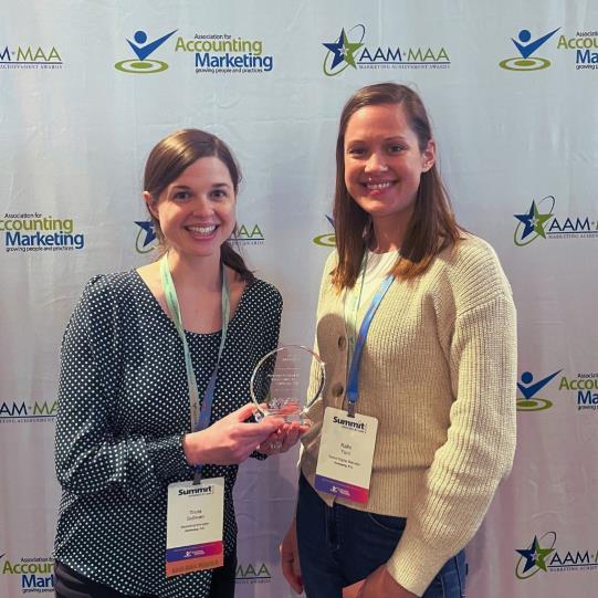 Two women in lanyards smile with a glass award in front of a white background patterned with the AAM MAA logo