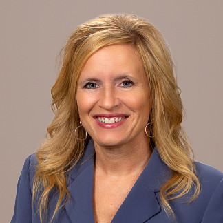 Woman with long blonde hair and a blue blazer smiles in front of a brown background