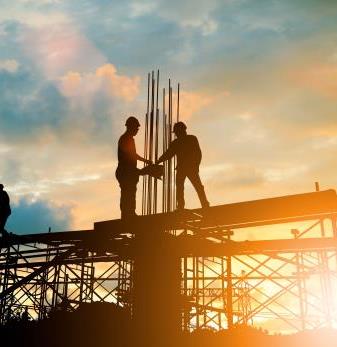 Silhouettes of two men in hard hats standing high up on a building frame with the sun behind them