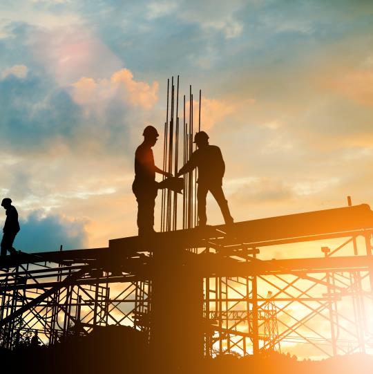 Silhouettes of two men in hard hats standing high up on a building frame with the sun behind them
