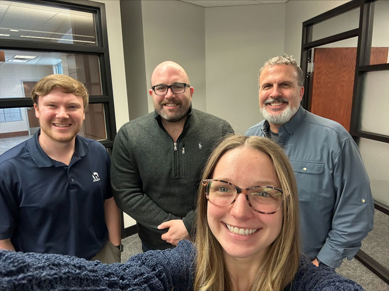 Three men and a woman take a selfie in an office space