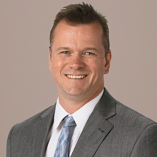 Dark haired man in a gray blazer smiles on a white background