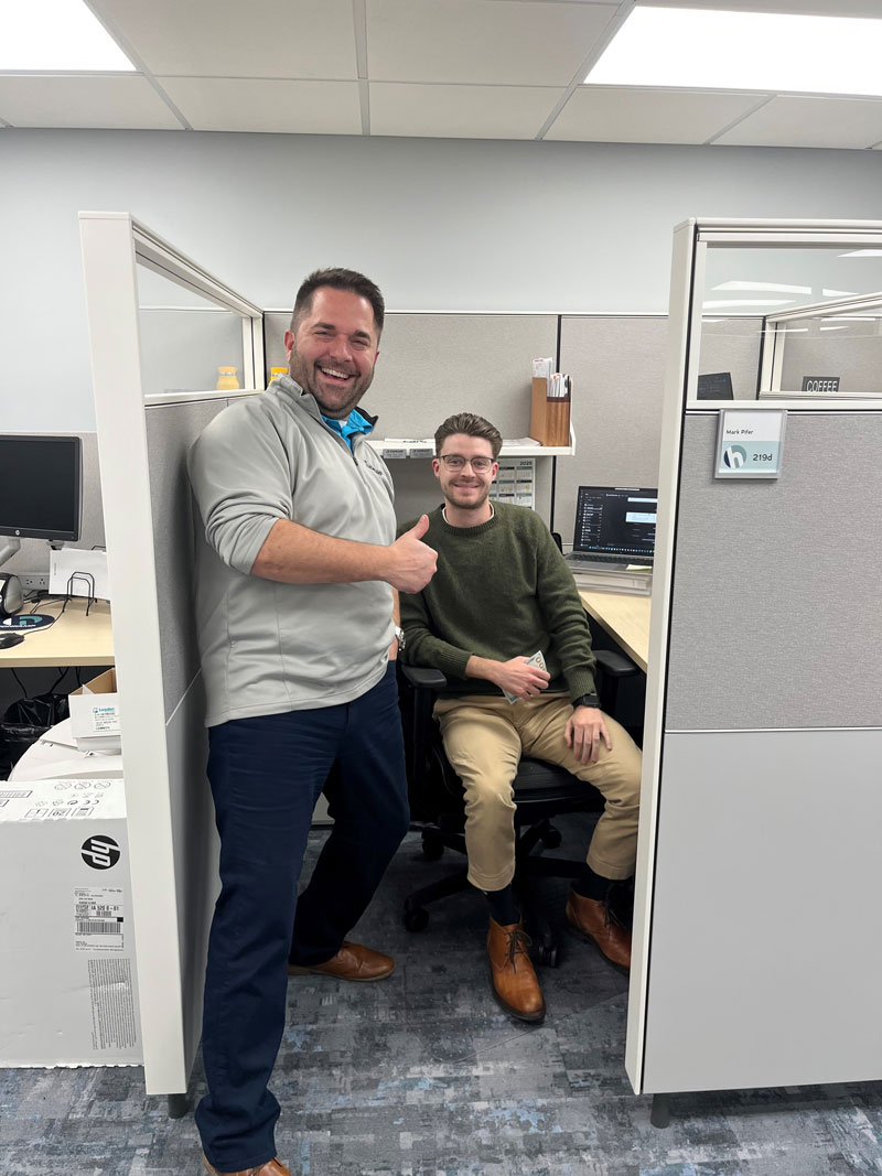 Two men smile and pose together in a cubicle in an office