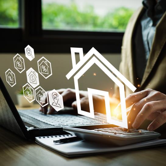 Close up of someone using a calculator and laptop as digital house icons float in the air