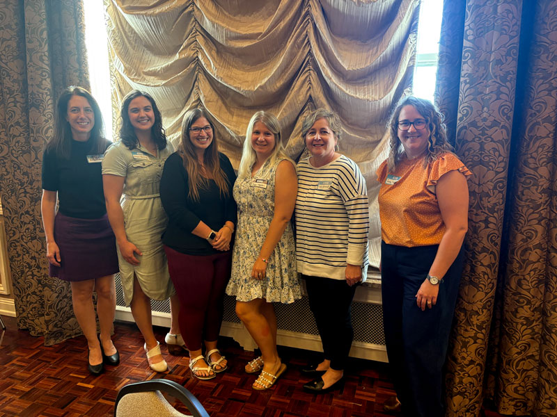 Six women in business clothes smile together in front of a curtained window