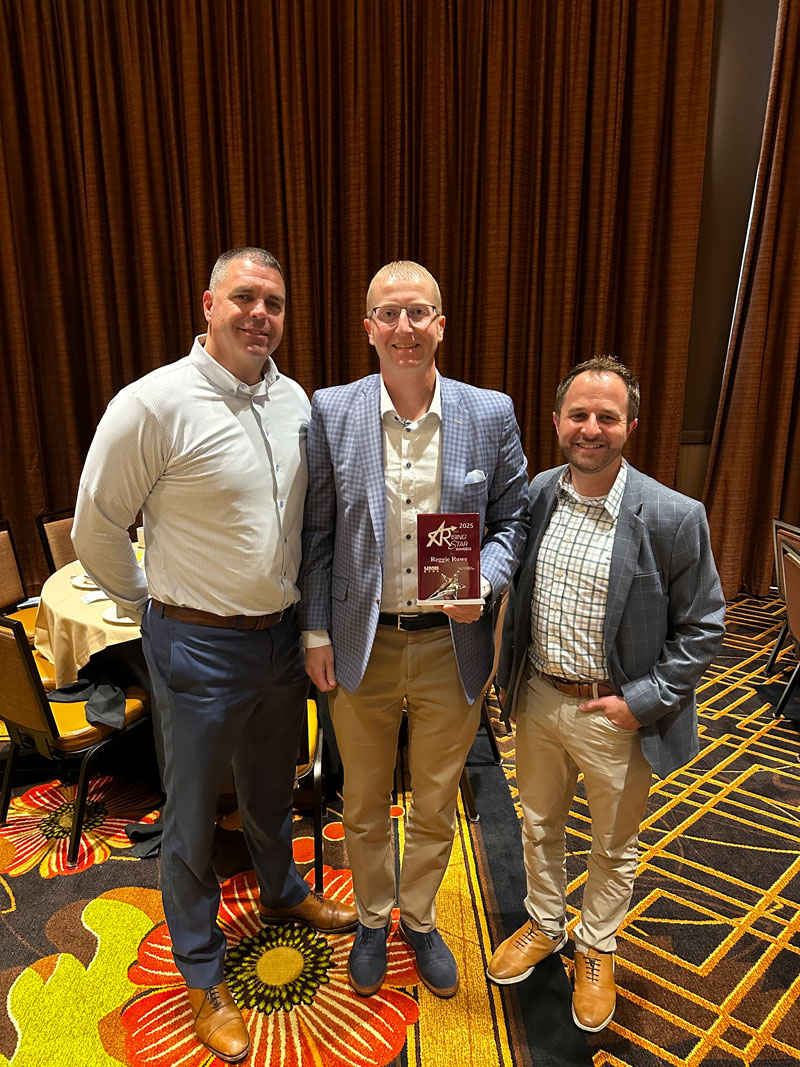 Three men in business clothes pose together with an award in an event space with colorful carpeting