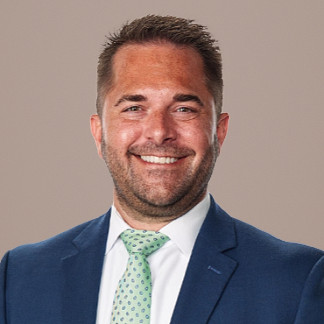 Dark haired man with a scruffy beard and navy blazer smiles on a white background
