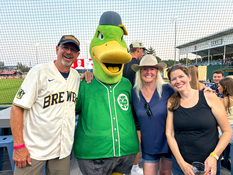 Two women and a man pose with a duck sports mascot at a baseball game