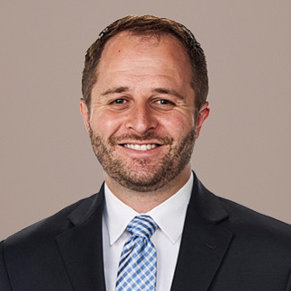Dark haired, bearded man in a black blazer smiles on a white background