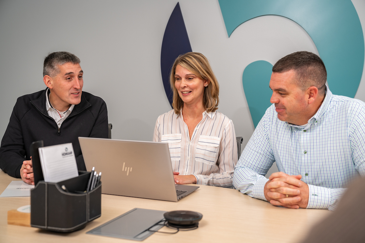 Two men and a woman sit in a conference room in front of a big sign with the lowercase h logo