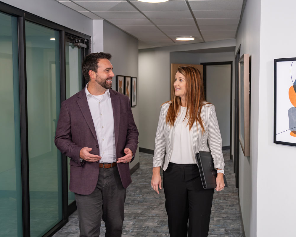 Man and woman in business clothes walk down a carpeted hallway
