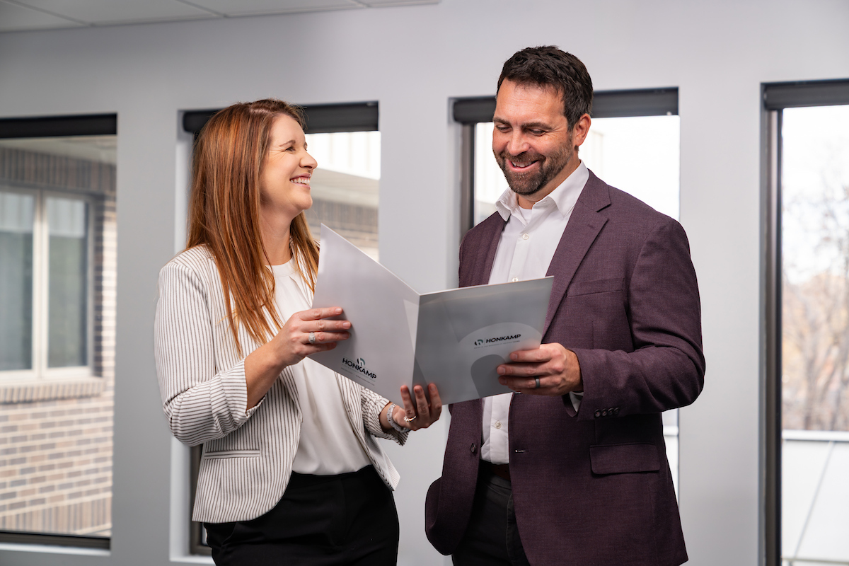 Man and a woman smile and look at a Honkamp folder in front of a row of bright windows