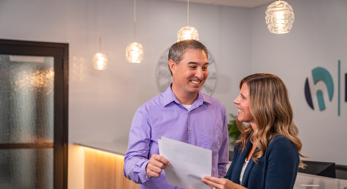 A man and woman laugh and hold papers in front of a reception desk in a Honkamp lobby