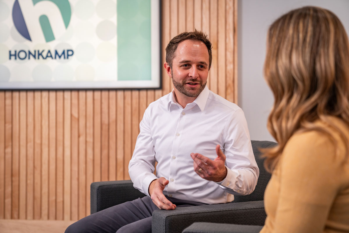 A man in a button down sits in a chair in front of a Honkamp sign and speaks to a woman obscured in the foreground