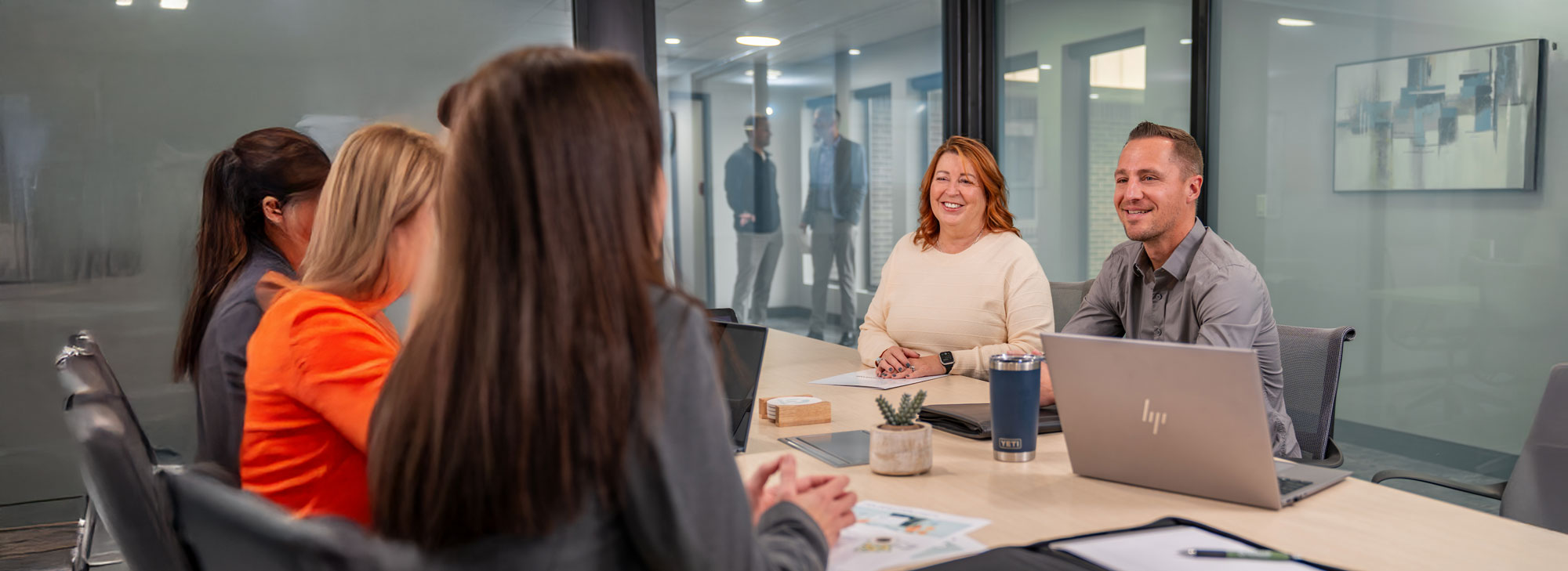 In a glass-walled conference room, a man and woman face a row of three women whose backs are to the camera