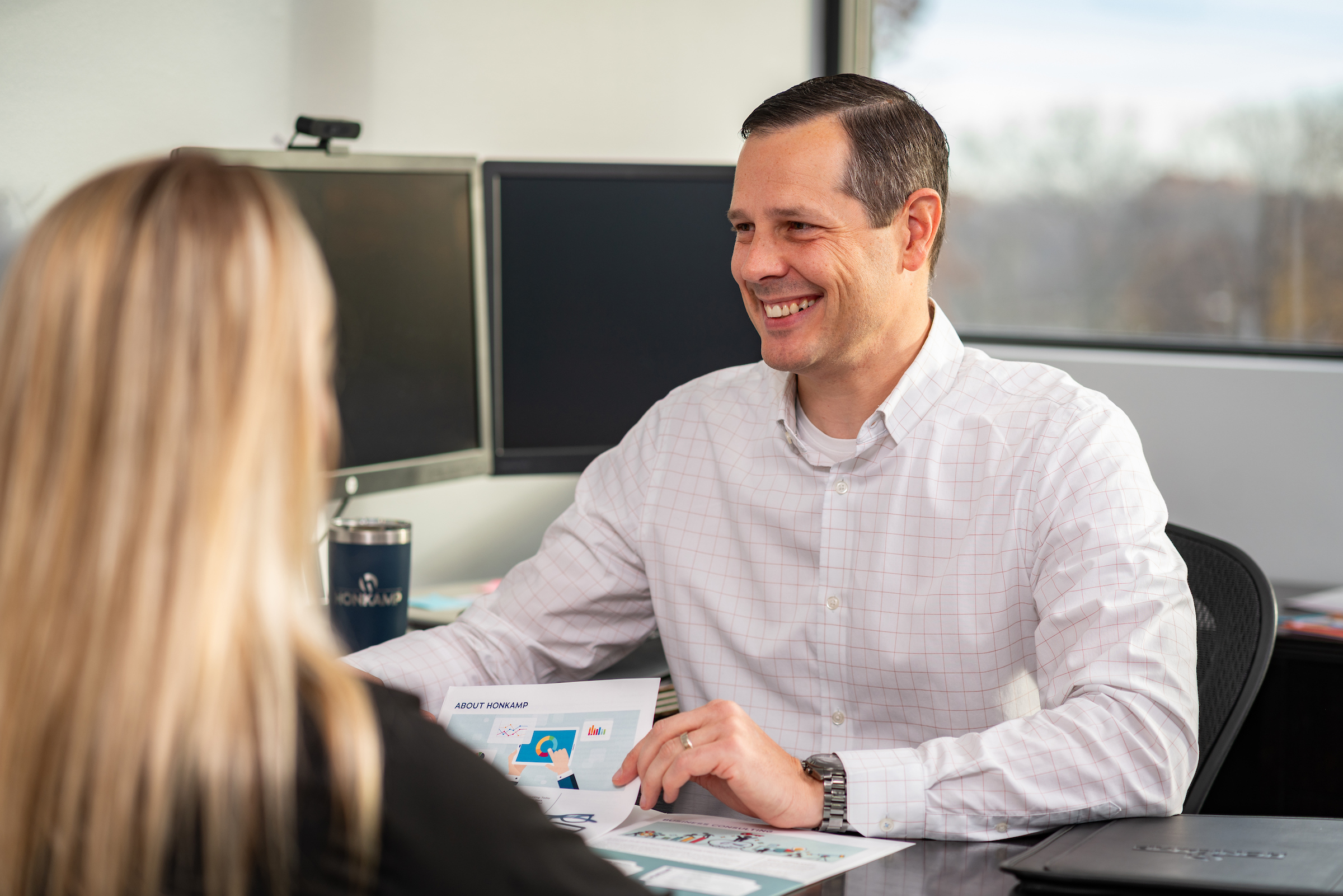 In a bright office, a man in a button down smiles at a blonde woman obscured in the foreground