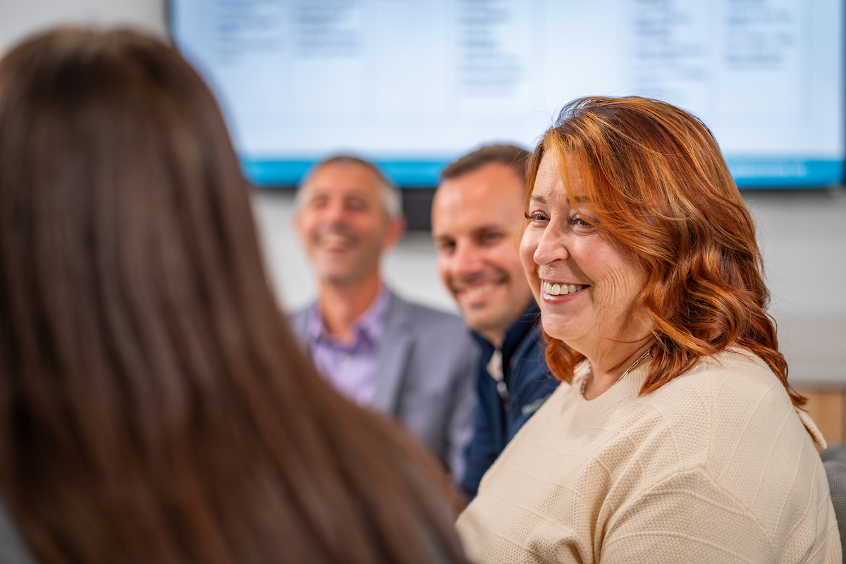 A red haired woman in a cream sweater smiles in a conference room with blurred coworkers behind her