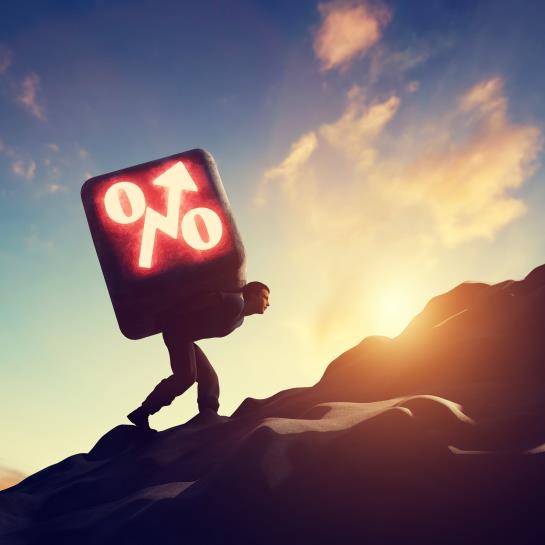 A man is hunched over and walking up a mountain, carrying a giant square on his back with a stock market symbol on it