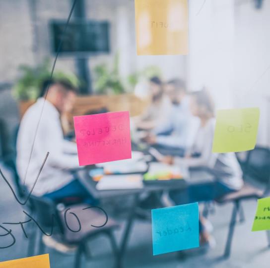 Post its are stuck to the opposite side of a glass wall, beyond which blurry office workers sit in a conference room