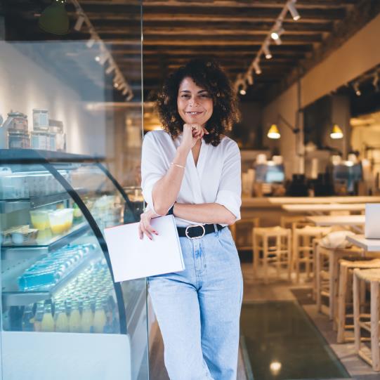 Curly haired woman leans against a bake case in a cafe space and holds papers, smiling into the camera