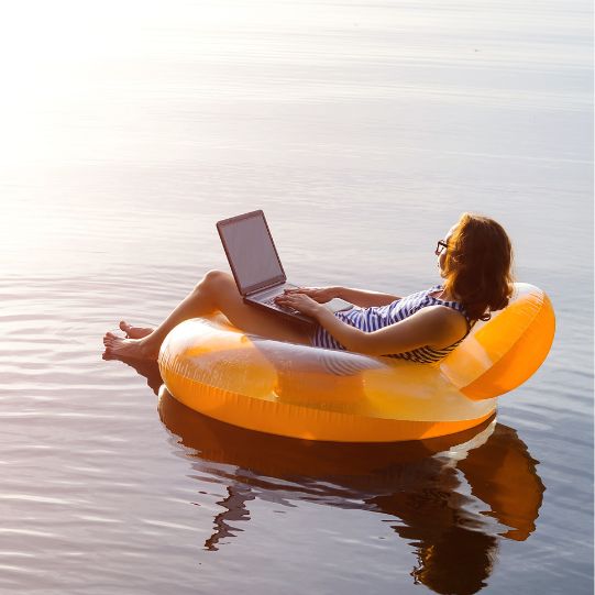 A woman in a bathing suit holds a laptop and floats in an orange tube on a body of water